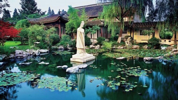 Een rustgevende tuin met een standbeeld, traditionele architectuur en een vijver met waterlelies.