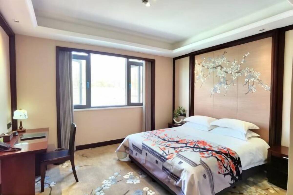 Ein Hotelzimmer mit einem großen Bett, einem Schreibtisch mit Lampe, einem Sessel und einem Fenster mit Vorhängen.