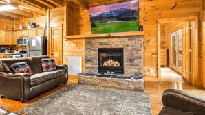 A cozy cabin interior with a stone fireplace, a leather sofa, and a mounted TV displaying a mountain landscape. - Fall Break Special!•Hot Tub•Game Room•Sleeps 22•3600 sq ft •Pigeon Forge (Pigeon Forge)