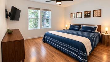A bedroom with a bed, wooden floor, ceiling fan, wall-mounted TV, and windows.
