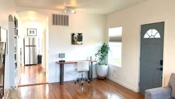A spacious living area with hardwood flooring, a white couch, a desk with a chair, a potted plant, and a grey door.