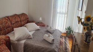 Makuuhuoneessa on suuri sänky, kuviollinen sohva, lamppu, ikkuna, jossa on ohuet verhot, ja maljakko, jossa on auringonkukkia.