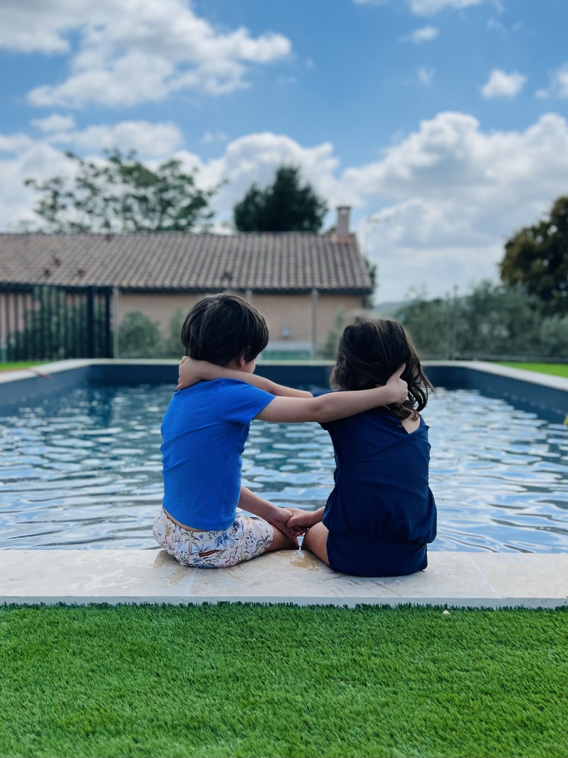 Two children sitting by a swimming pool with a house in the background.