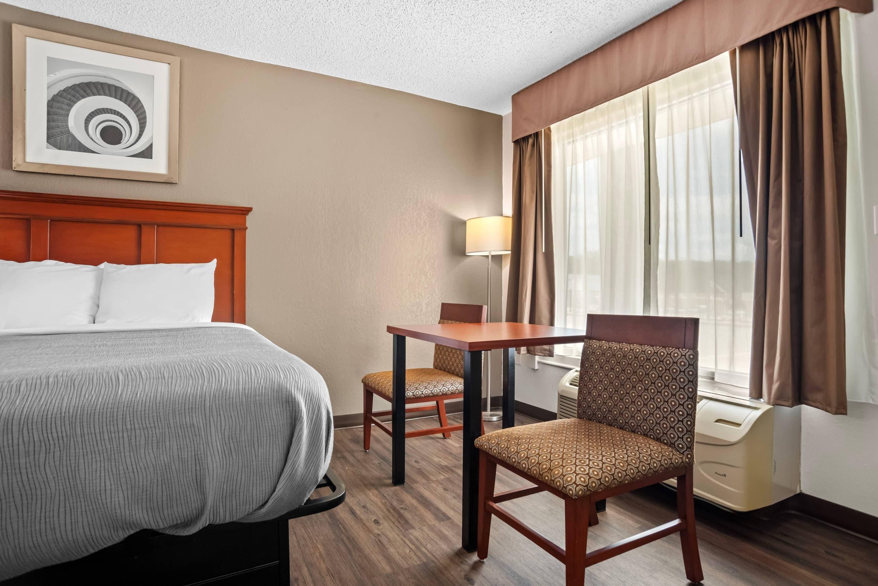 Una habitación de hotel con una cama, un escritorio, dos sillas, una lámpara y una ventana con cortinas.