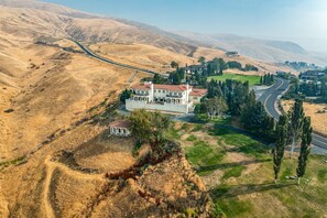 Une grande maison blanche à plusieurs étages, avec un toit recouvert de tuiles rouges, entourée d’herbe sèche et de collines.