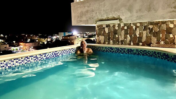 An outdoor swimming pool with a tiled edge, a stone wall, and a view of buildings in the background. - Hotel María Bonita Higüey,  room 301 Family property centrally located (Higüey)