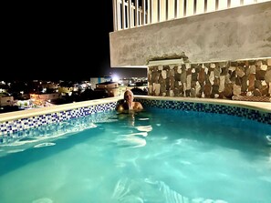 Una piscina al aire libre con borde de azulejos, pared de piedra y vista a edificios al fondo.