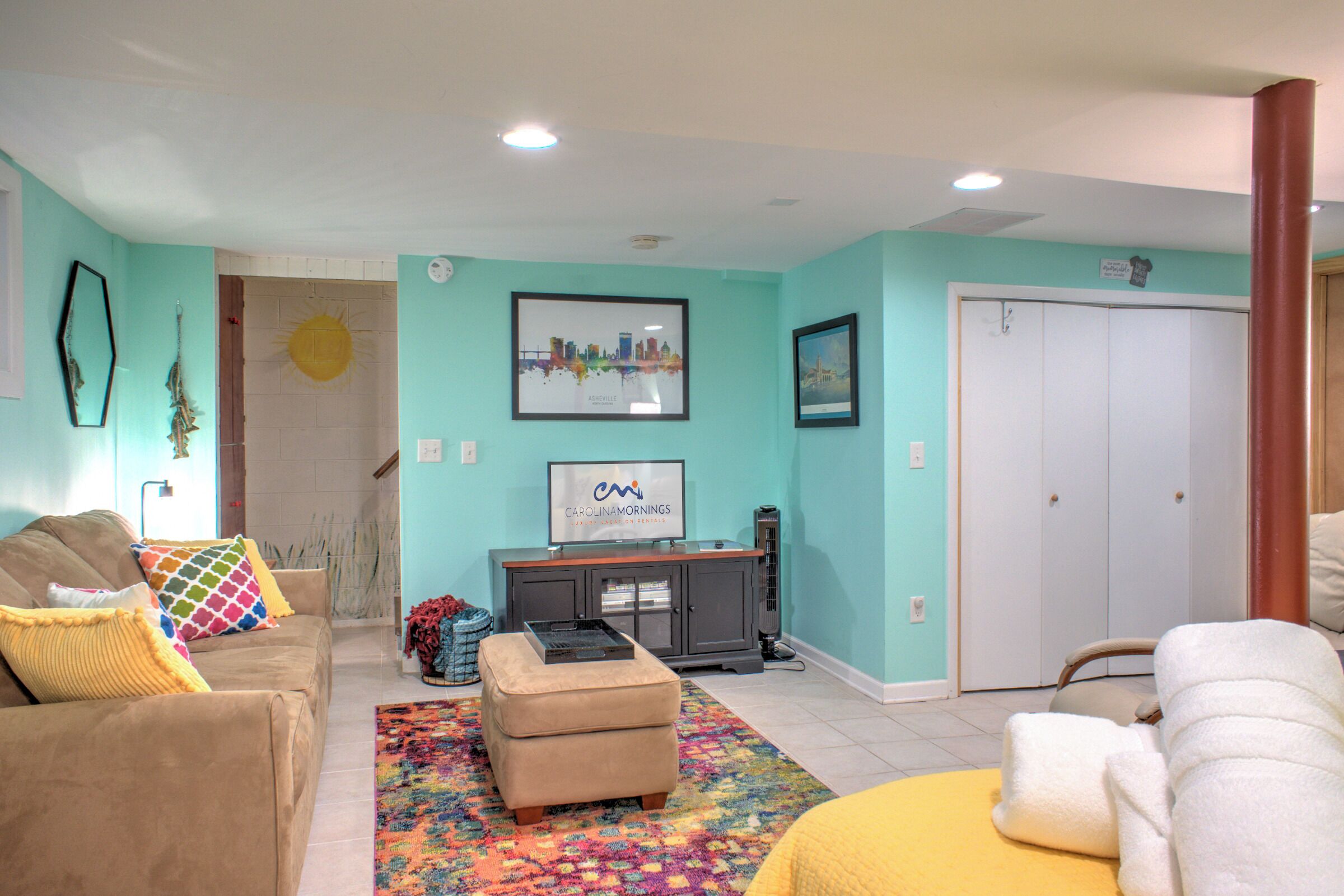 A living room with a sofa, ottoman, and a colorful rug. There is a television on a stand, a door, and a closet in the background.