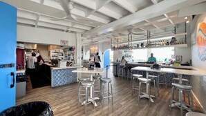 Un bar moderne avec de hauts plafonds et des poutres apparentes, doté d’un comptoir avec des tabourets et d’une variété de bouteilles exposées sur des étagères.