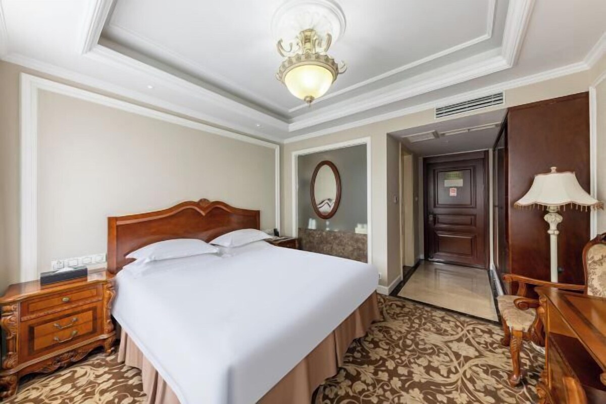 Quarto de hotel com uma cama grande, mesa de cabeceira de madeira e uma escrivaninha com abajur.