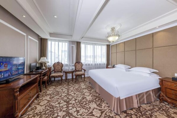 Quarto de hotel com uma cama grande, televisão, escrivaninha com abajur, duas cadeiras e um tapete estampado.