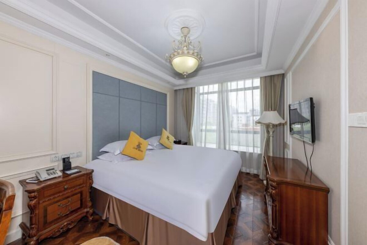 Quarto de hotel com uma cama grande, cômoda de madeira, mesa de cabeceira com telefone e televisão na parede.