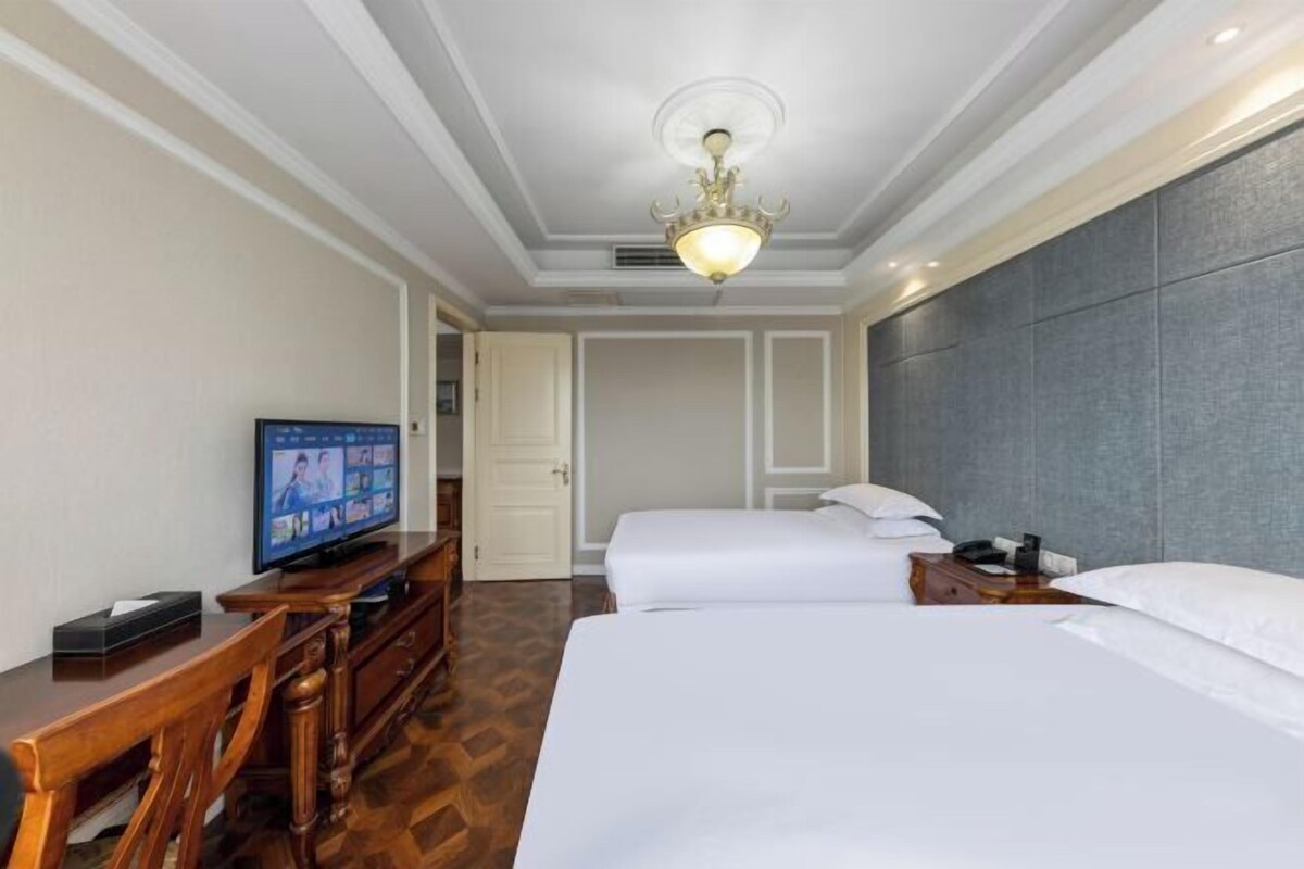 Quarto de hotel com televisão, duas camas, escrivaninha de madeira e um candelabro.