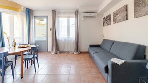A living room with a grey sofa, a dining table with chairs, and a window with curtains. - Peaceful Apartment Surrounded by Nature – Quiet Location Minutes from Assisi (Pian della Pieve)