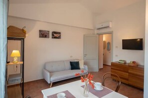 Quarto pequeno e bem iluminado, com sofá, mesa de jantar com um vaso de flores, televisão na parede e um armário de madeira.