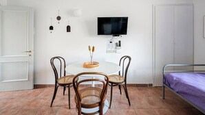 A small, clean room with a round dining table, two wicker chairs, a bed with a purple bedspread, a TV mounted on the wall, and a door with a gold handle.