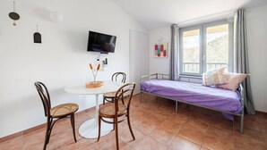 A small, tidy room with a bed, a TV, a dining table with chairs, and a window with curtains.