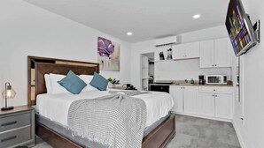 A modern bedroom with a large bed, bedside tables, a flat-screen TV, and a kitchenette with white cabinets and appliances. - Charming Studio Walking Distance To Ocean - Unit 8 (Hollywood)