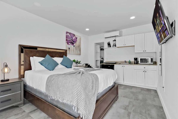A modern bedroom with a large bed, bedside tables, a flat-screen TV, and a kitchenette with white cabinets and appliances.