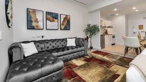 Un salon moderne avec un canapé en cuir noir, un coin repas avec une chaise blanche et une cuisine en arrière-plan.
