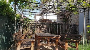 Un banc et une table en bois sont placés sous une pergola couverte de plantes grimpantes, le tout étant entouré d’une clôture en bois et d’un mur de pierre.