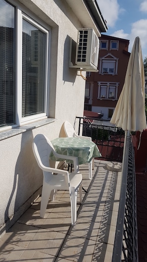 Un balcón con dos sillas de plástico blanco y una mesa, un equipo de aire acondicionado y una reja metálica.