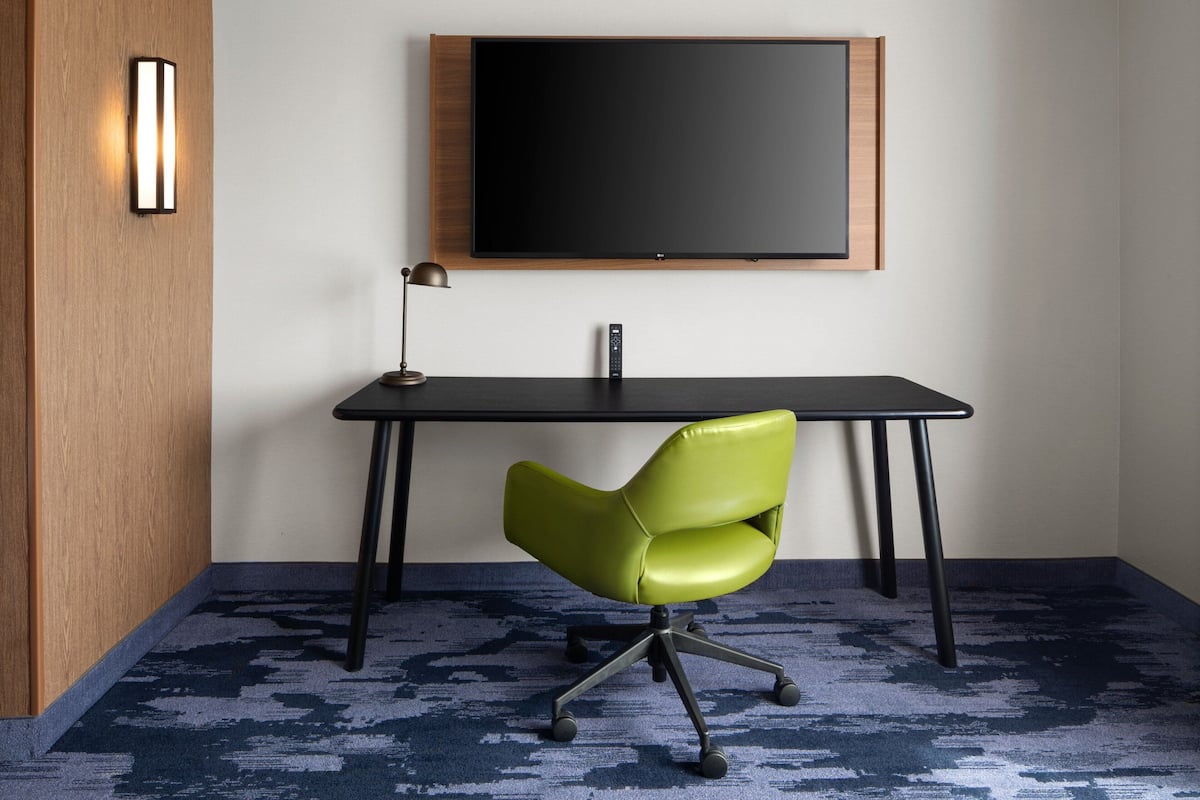 Een kamer met een houten wand, een zwart bureau, een groene bureaustoel, een zwarte lamp en een groot zwart scherm dat aan de muur is bevestigd.