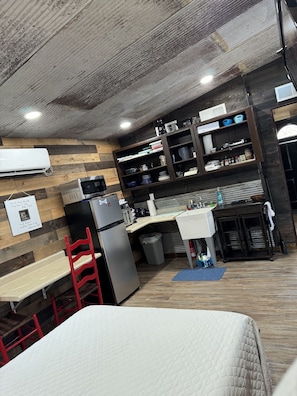 A compact kitchen with a refrigerator, microwave, and sink. There is a small dining table and a red chair. The kitchen has wooden paneling on the walls and a ceiling with exposed beams. - The Ridge Adventure Center rustic pet friendly tiny cabin, outdoor shower stage  (Jane)