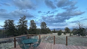 Une terrasse en bois avec une table et des chaises en verre, entourée d’arbres et offrant une vue sur un paysage montagneux.