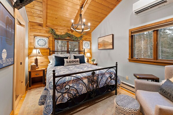 A cozy bedroom with a wooden ceiling, a bed with a patterned bedspread, two bedside lamps, a nightstand, a wicker chair, and a window with a view of trees.