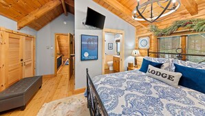 Une chambre confortable avec un plafond en bois, un lit recouvert d’une courtepointe à motifs bleus et blancs, un oreiller orné du mot « Lodge » et un téléviseur fixé au mur.