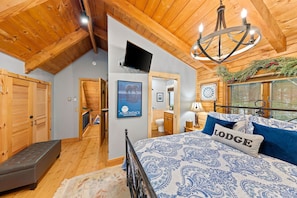 A cozy bedroom with a wooden ceiling, a bed with a blue and white patterned bedspread, a pillow with the word "Lodge," and a television mounted on the wall.