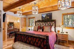 A cozy bedroom with a wooden ceiling, a bed with a red and black patterned bedspread, a wooden nightstand, and a window with a view of trees.