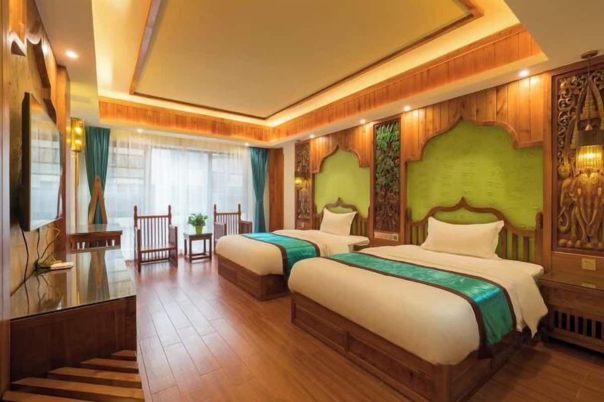 A hotel room with two beds, wooden flooring, a desk with a chair, and a large window with curtains.