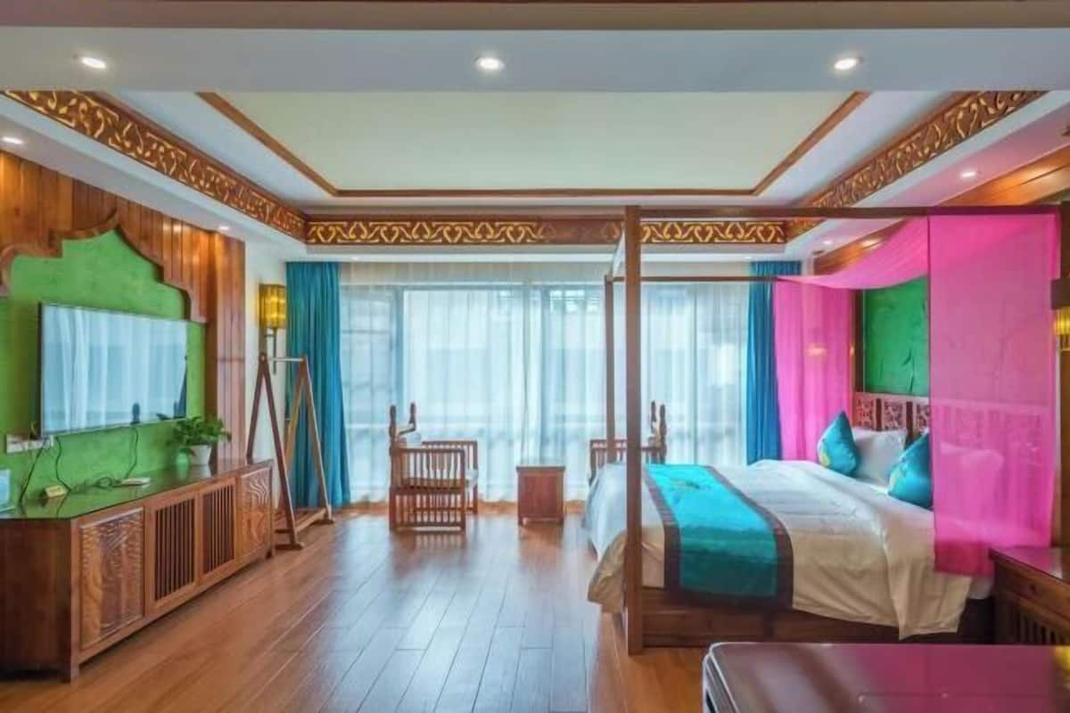 A hotel room with a four-poster bed, wooden flooring, a green accent wall, and a large window with curtains.