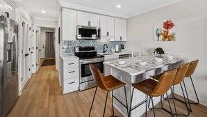 A modern kitchen with white cabinets, a stainless steel refrigerator, a microwave, and a dining area with a white island and brown chairs.