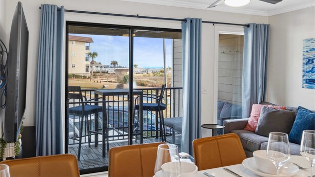 A room with a balcony, a dining table set with plates and glasses, a sofa, and a ceiling fan.