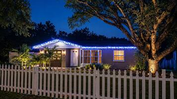 Une maison avec une clôture en bois peinte en blanc, un arbre au premier plan et des guirlandes lumineuses sur le toit.