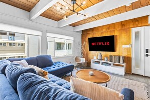 A modern living room with a blue sofa, a wooden coffee table, and a mounted TV displaying 'NETFLIX'. - Walk to Beach & Bay | Family Friendly (San Diego)