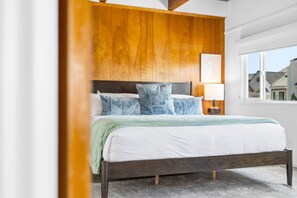 A bedroom with a wooden headboard, a bed with white bedding and blue pillows, a bedside lamp, and a view of buildings through a window.