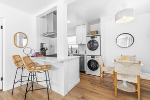 A modern kitchen with white cabinetry, a central island with bar stools, a built-in washing machine, and a dining area with a round table and chairs.
