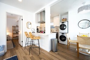 Una cocina compacta con lavadora, dos taburetes, un cubo de basura y una zona de comedor con mesa y sillas.