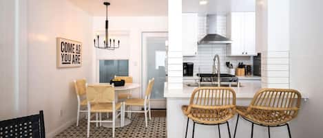 A modern kitchen with white cabinetry, a black stove, and a stainless steel sink. There are two wicker chairs and a dining table with chairs.