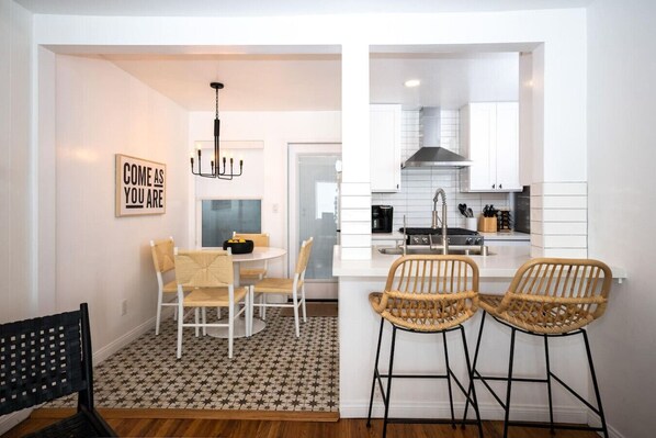 A modern kitchen with white cabinetry, a black stove, and a stainless steel sink. There are two wicker chairs and a dining table with chairs.
