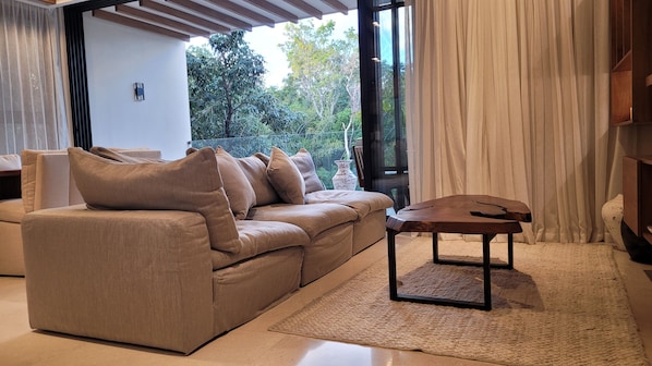 A modern living room with a beige sofa, a wooden coffee table, and a view of greenery through large windows.