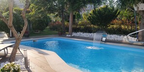 A swimming pool with a water feature, surrounded by a landscaped garden with trees and a stone wall. - Apartment in a house with garden, pool and barbecue, 10 minutes from Pontevedra (Vilaboa, Pontevedra)