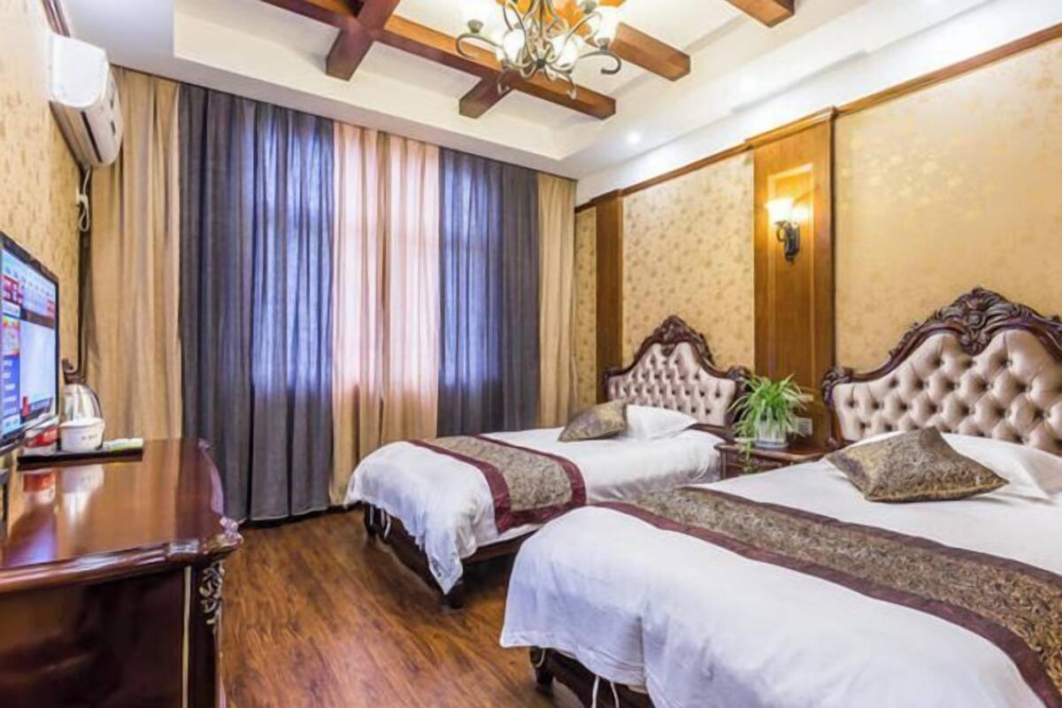 A hotel room with two beds, a wooden desk, a television, and a large window with curtains.