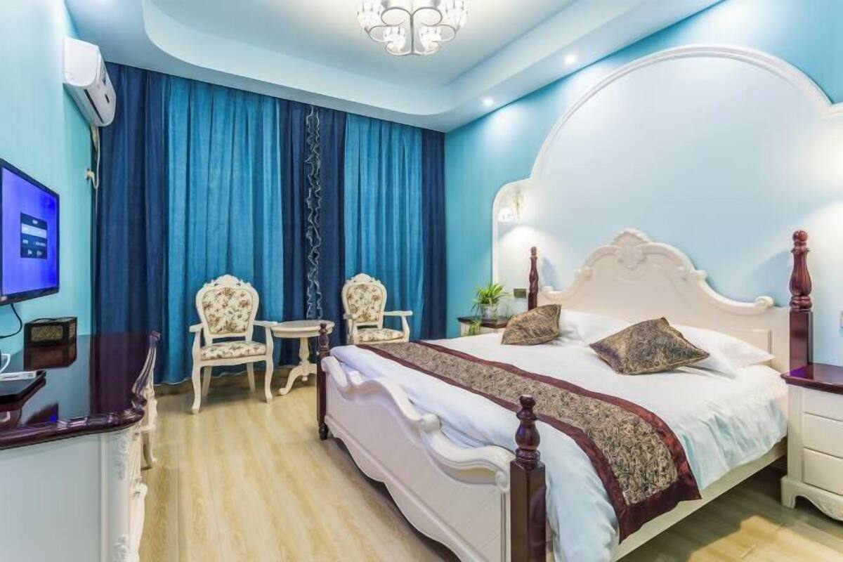 A bedroom with a large bed, two chairs, a small table, a TV, and a chandelier.