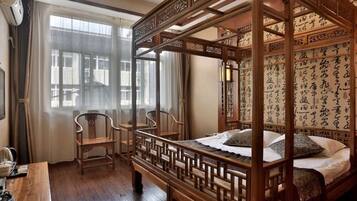 Una cama tradicional con dosel de madera y cuatro columnas, un escritorio con silla, y una ventana con cortinas.