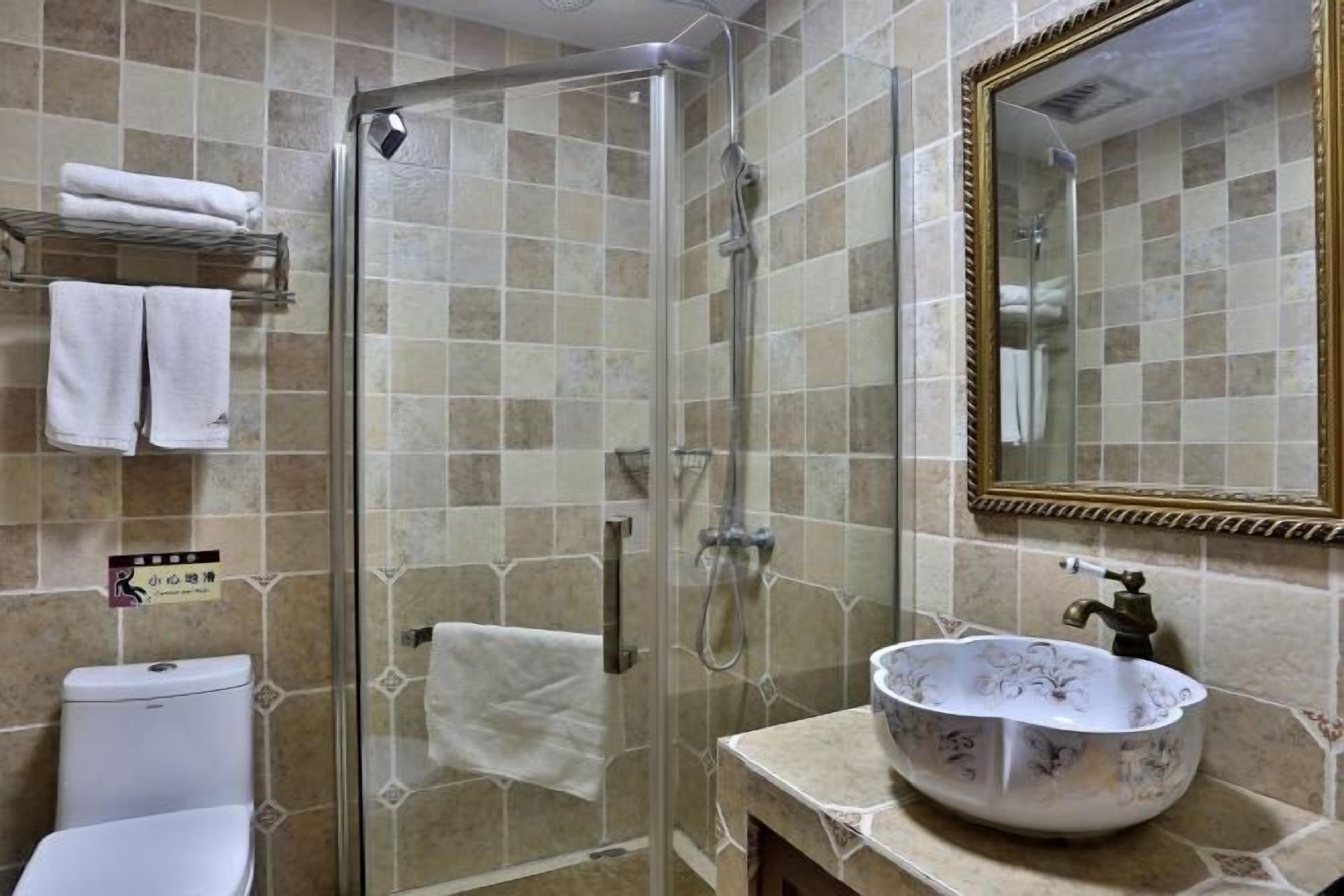 Un baño con ducha de vidrio, inodoro blanco, lavamanos con un recipiente decorativo y un espejo con marco dorado.